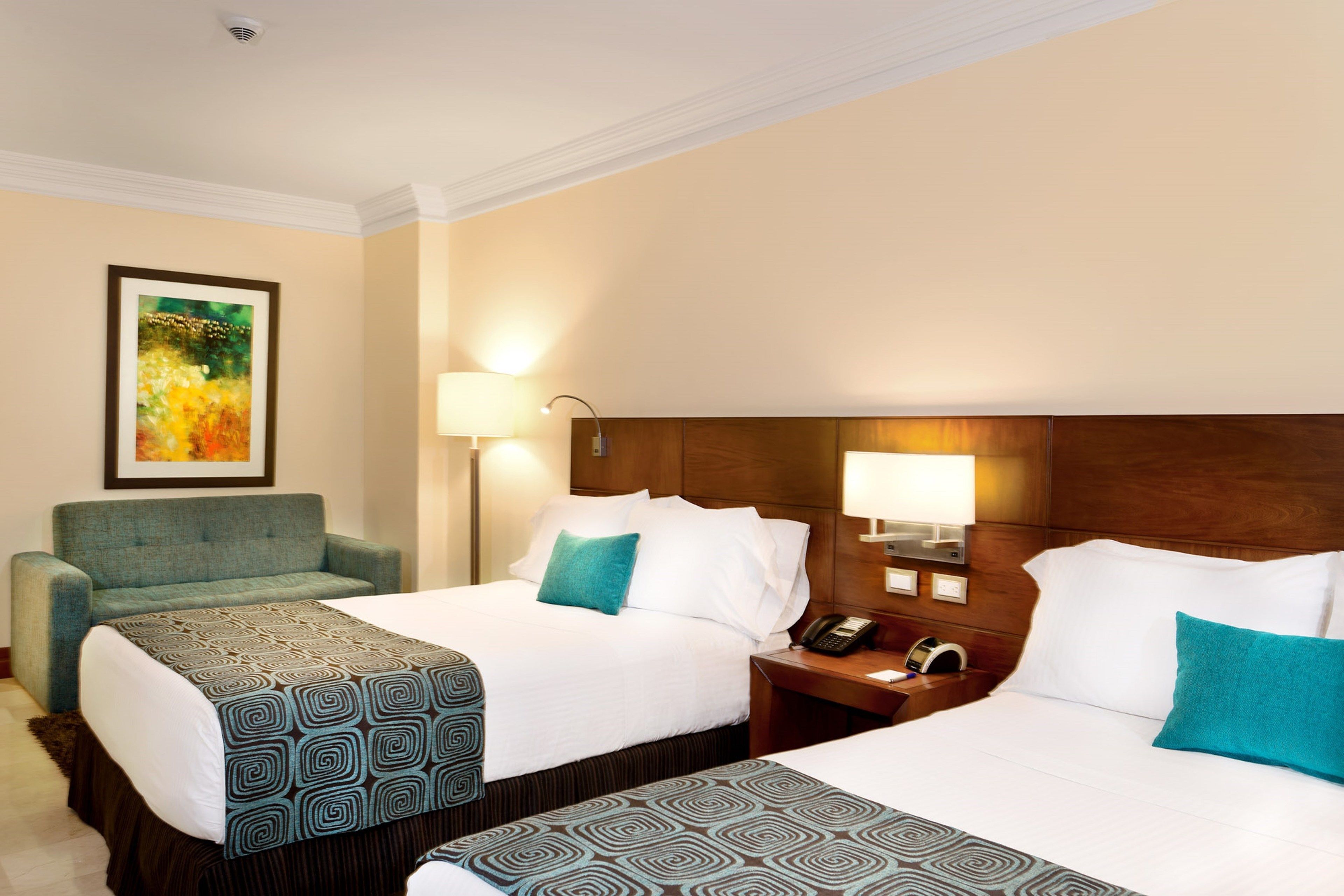 Ein Hotelzimmer mit zwei Betten, einem Sofa, einem Gemälde an der Wand und einem Nachttisch mit Lampe und Telefon.