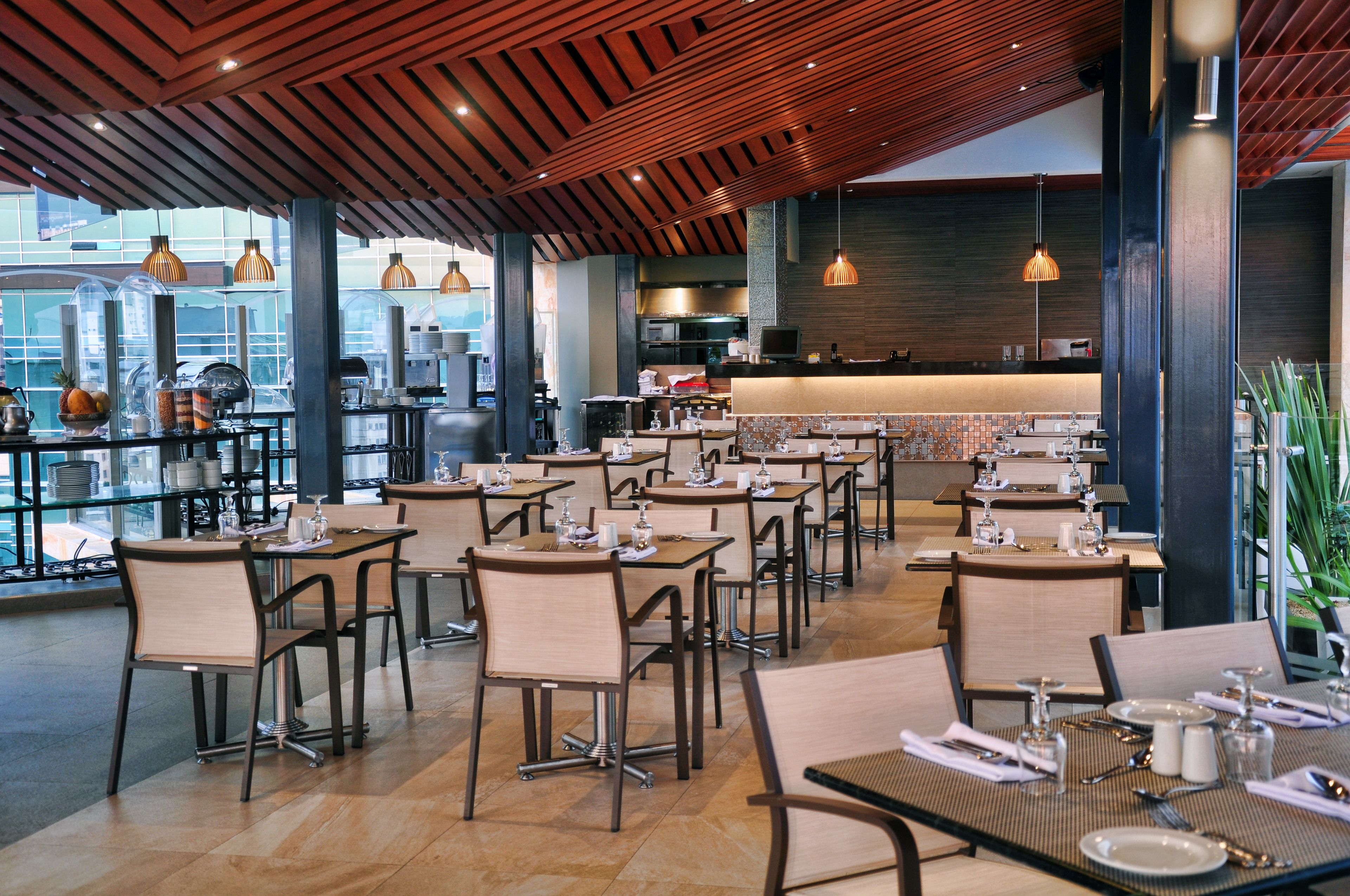 Un restaurante moderno con varias mesas y sillas, una zona de cocina y grandes ventanales con vistas a la ciudad.