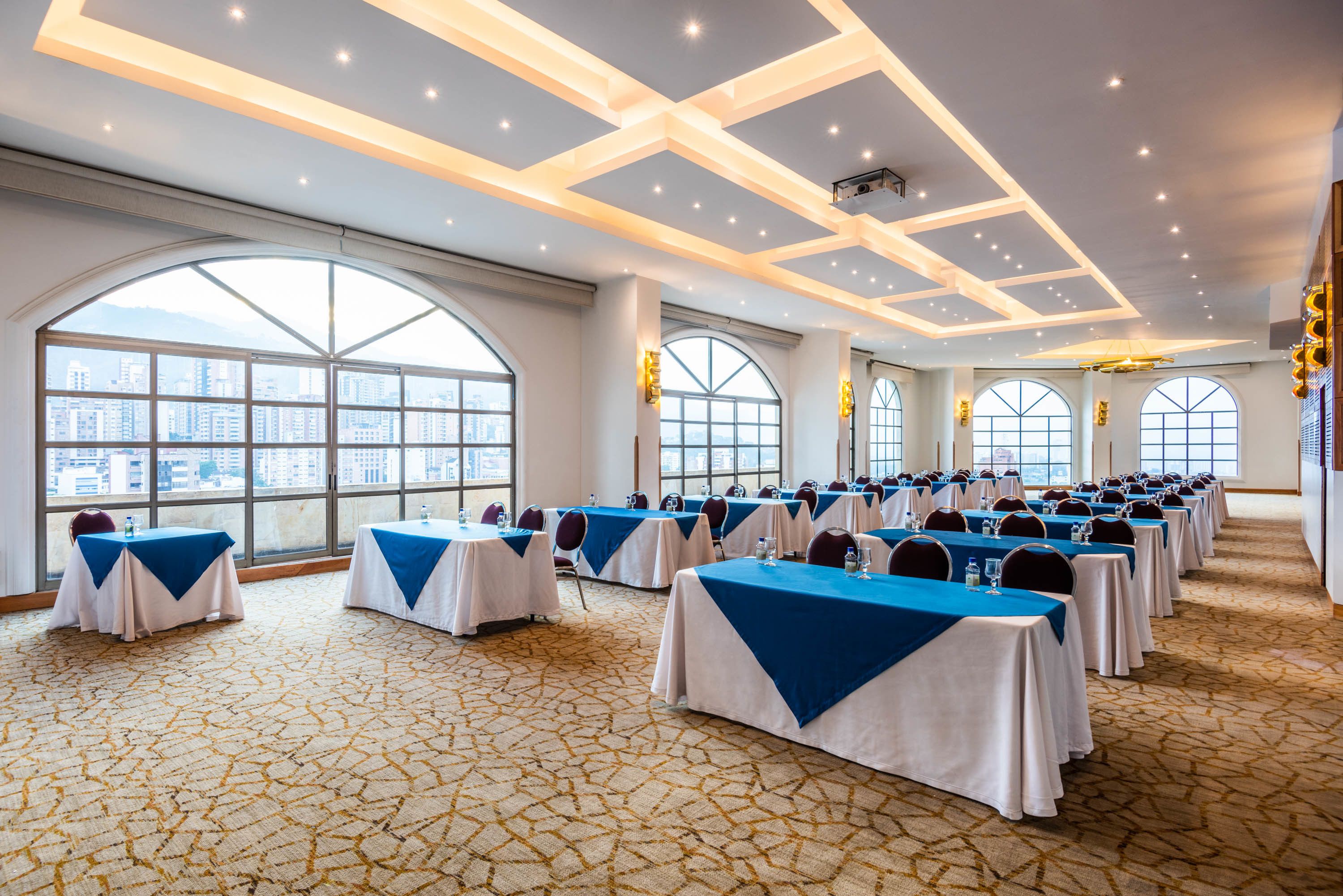 Un salón de conferencias con mesas dispuestas en filas, ventanas amplias con vistas a la ciudad y una alfombra estampada.