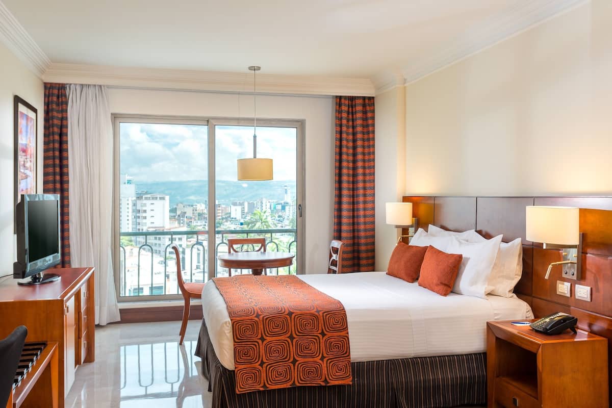 Ein Hotelzimmer mit Bett, Schreibtisch, Stuhl, Fernseher und einem Balkon mit Blick auf Häuser und die Stadtlandschaft.
