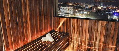 A wooden sauna with a bench and a towel, a window with a cityscape view at night.