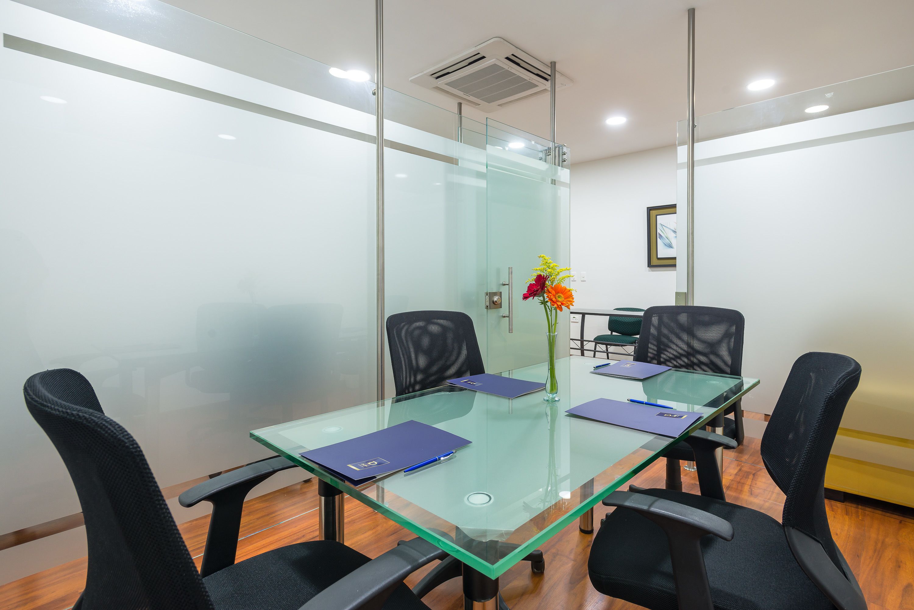 A conference room with a glass table, four black chairs, and a vase with flowers.