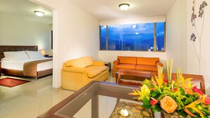 Una habitación de hotel con una cama, un sillón amarillo, un sofá naranja, una mesa de centro de vidrio y vistas a las montañas desde la ventana.