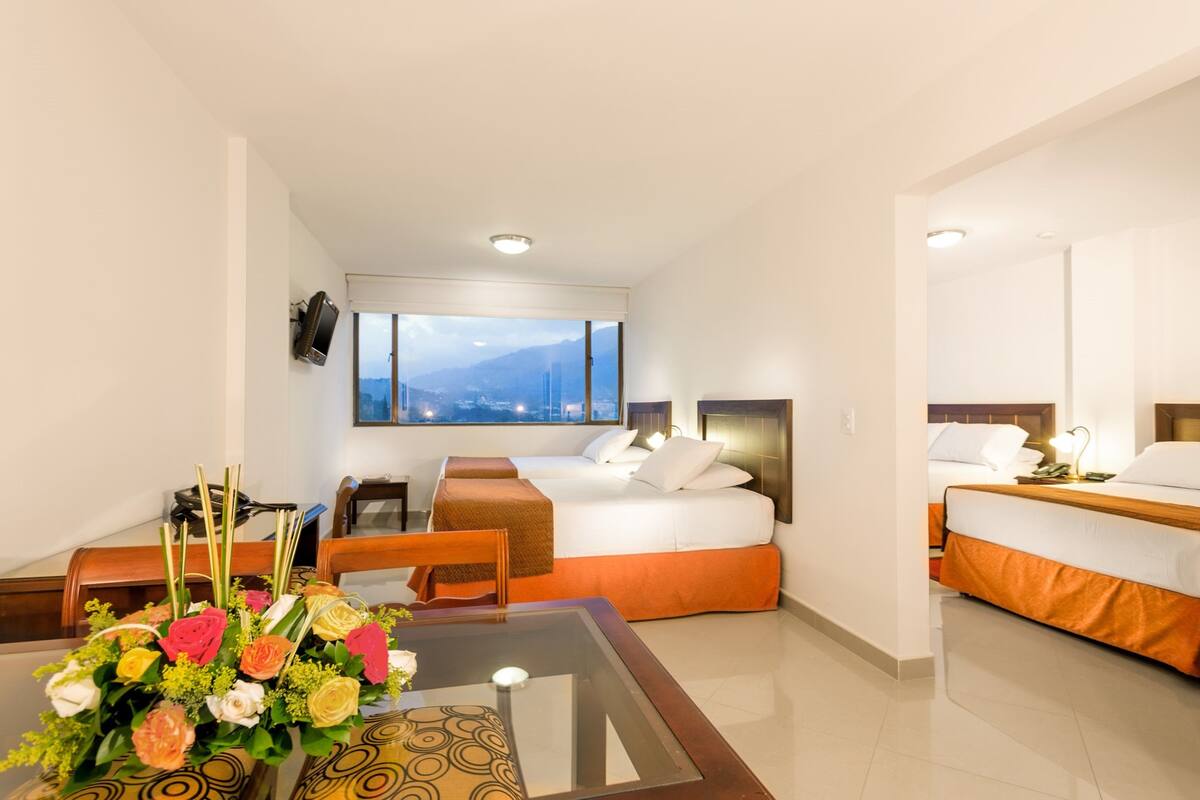 Una habitación de hotel con cama, escritorio, silla, mesa con un jarrón de flores y un gran ventanal con vistas a la montaña.