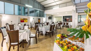 Un comedor amplio con varias mesas y sillas, un bufé con frutas y aperitivos variados, y un techo alto con ventanas grandes.