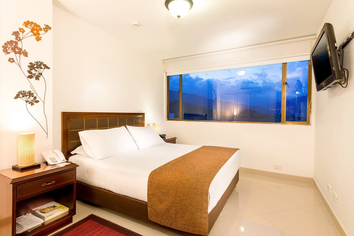 Habitación de hotel con cama, mesita de noche con lámpara, televisor montado en la pared y ventana con vista a la ciudad al anochecer.