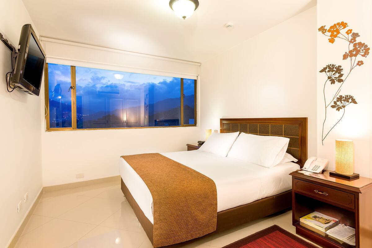 Habitación de hotel con una cama grande, una mesita de noche con lámpara, un televisor de pantalla plana en la pared y una ventana con vistas a la ciudad.