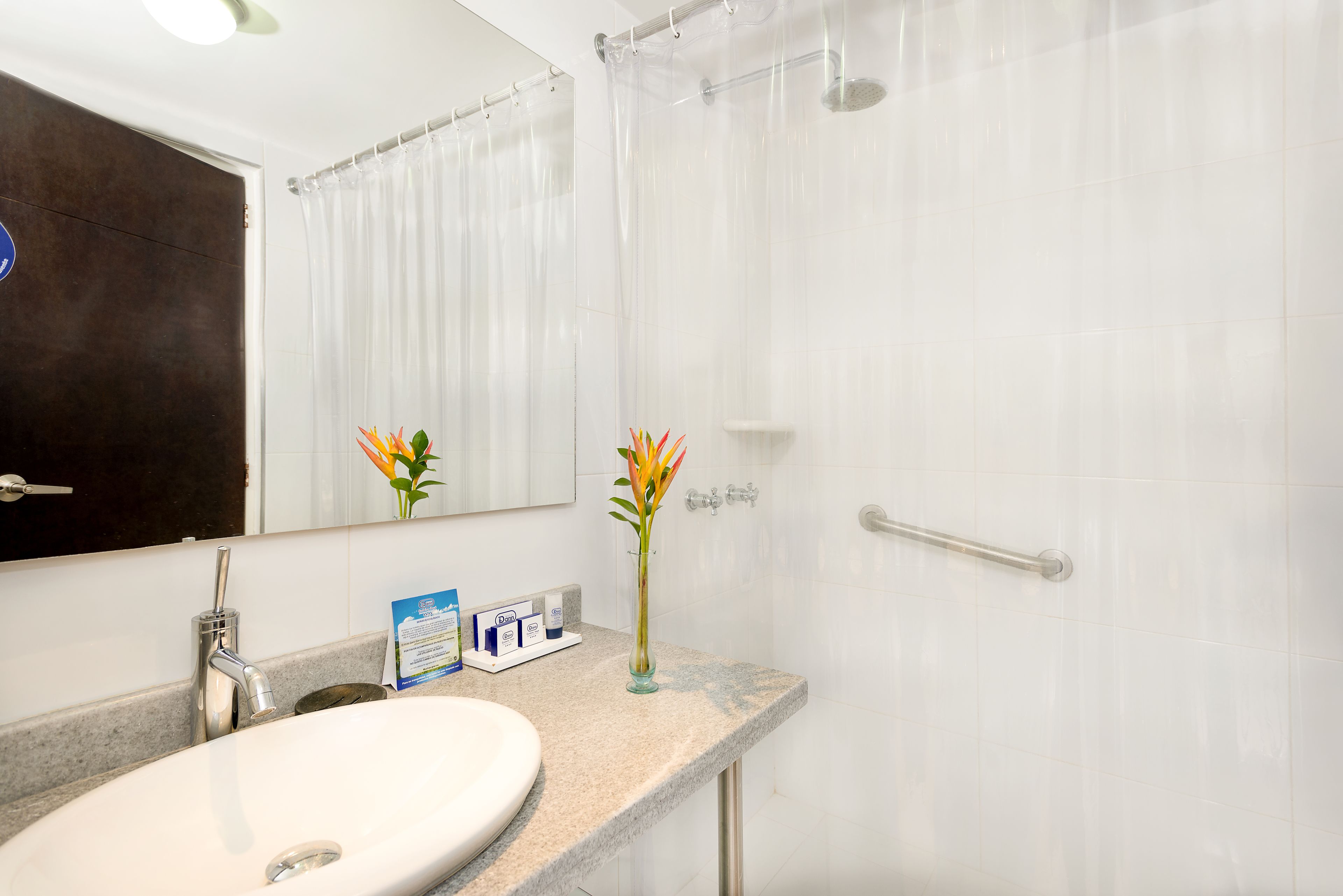 Une salle de bain avec un lavabo blanc, un miroir, un rideau de douche et un vase rempli de fleurs posé sur le comptoir.