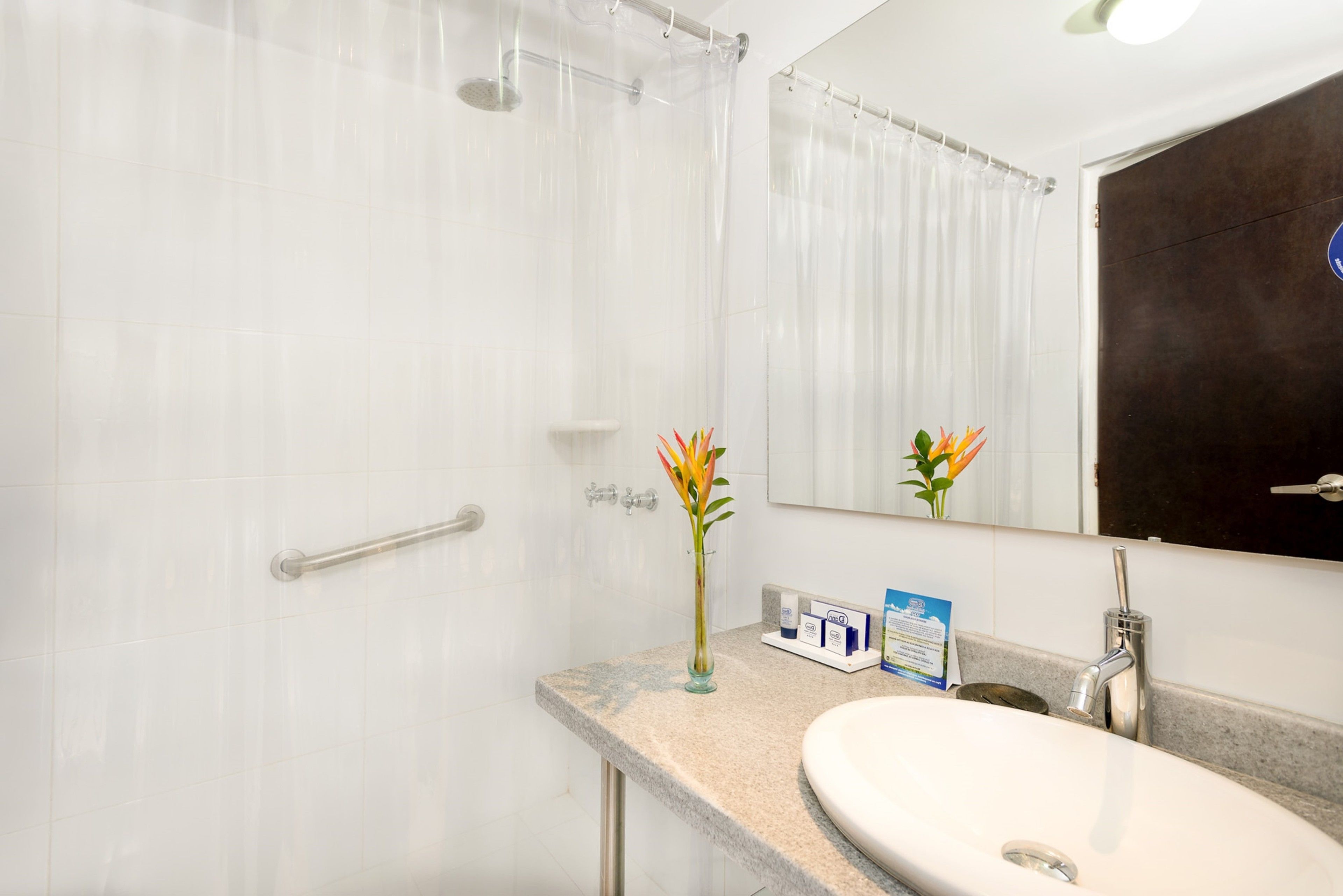 Un baño con ducha, lavamanos, espejo y un florero con flores.
