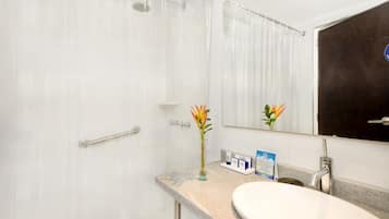 Un baño con ducha, lavamanos, espejo y un florero con flores.
