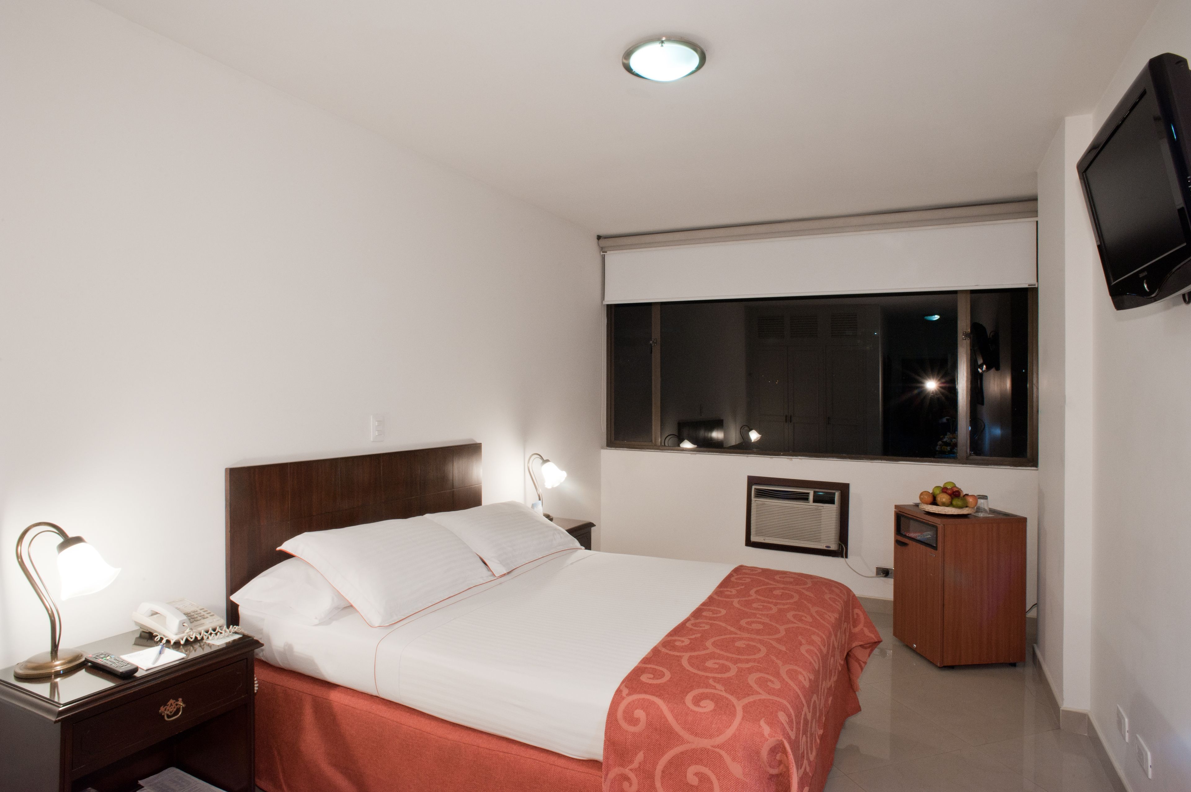 A hotel room with a bed, bedside lamp, nightstand, and a television mounted on the wall.