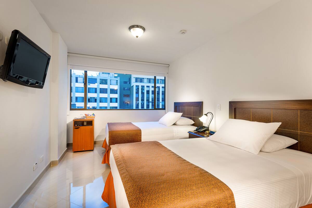 Habitación de hotel con dos camas, un televisor montado en la pared, una ventana con vistas a edificios y un pequeño armario de madera.