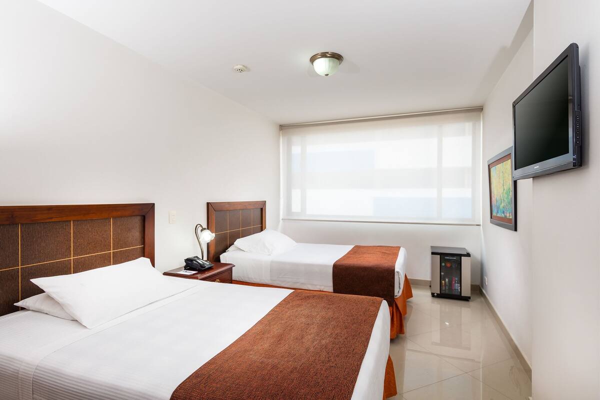 Habitación de hotel con dos camas, televisión, mesita de noche, lámpara, ventana y un minibar.