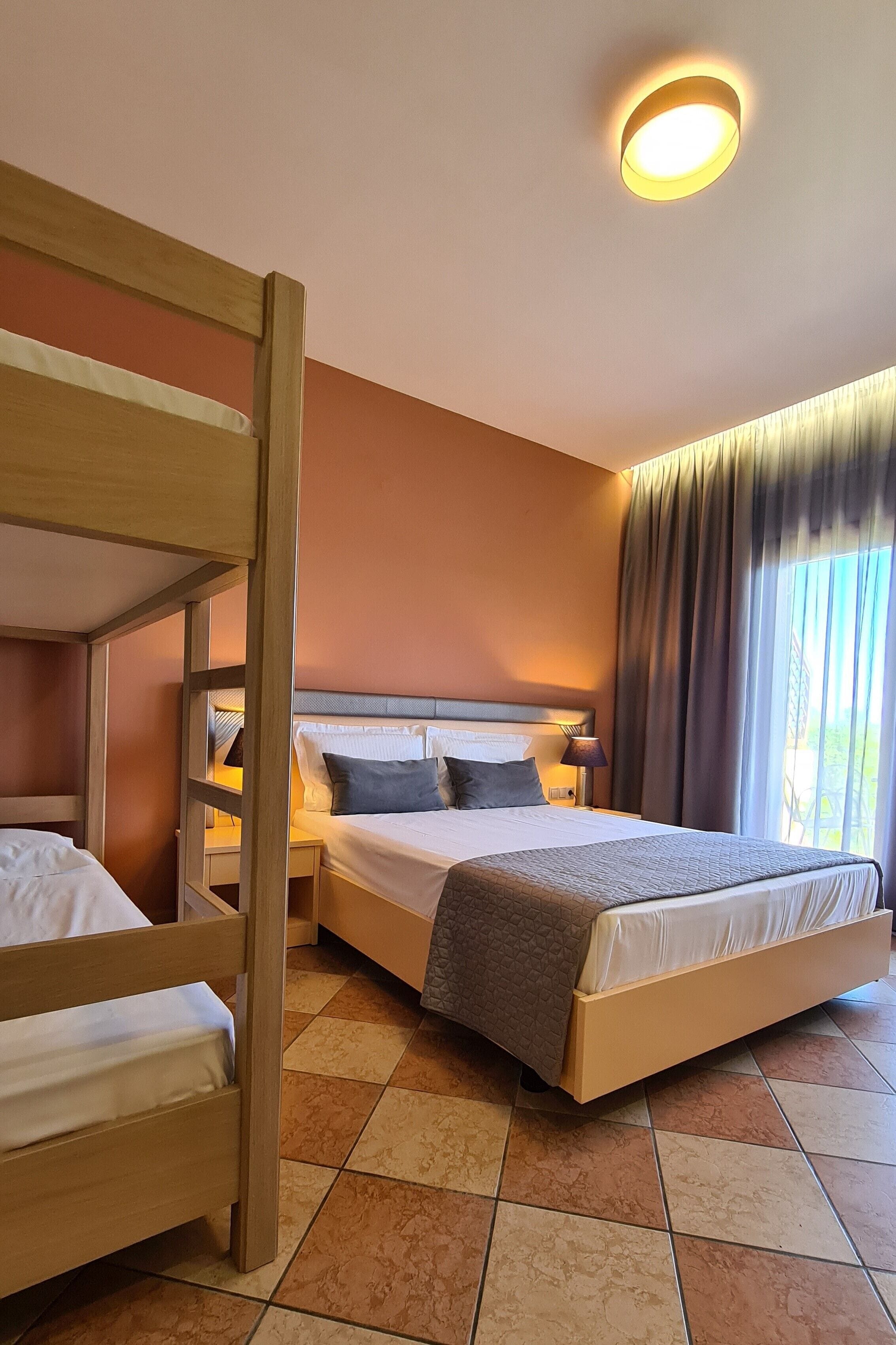 Habitación de hotel con literas, una cama individual, una mesita de noche con lámpara y un ventanal con cortinas.