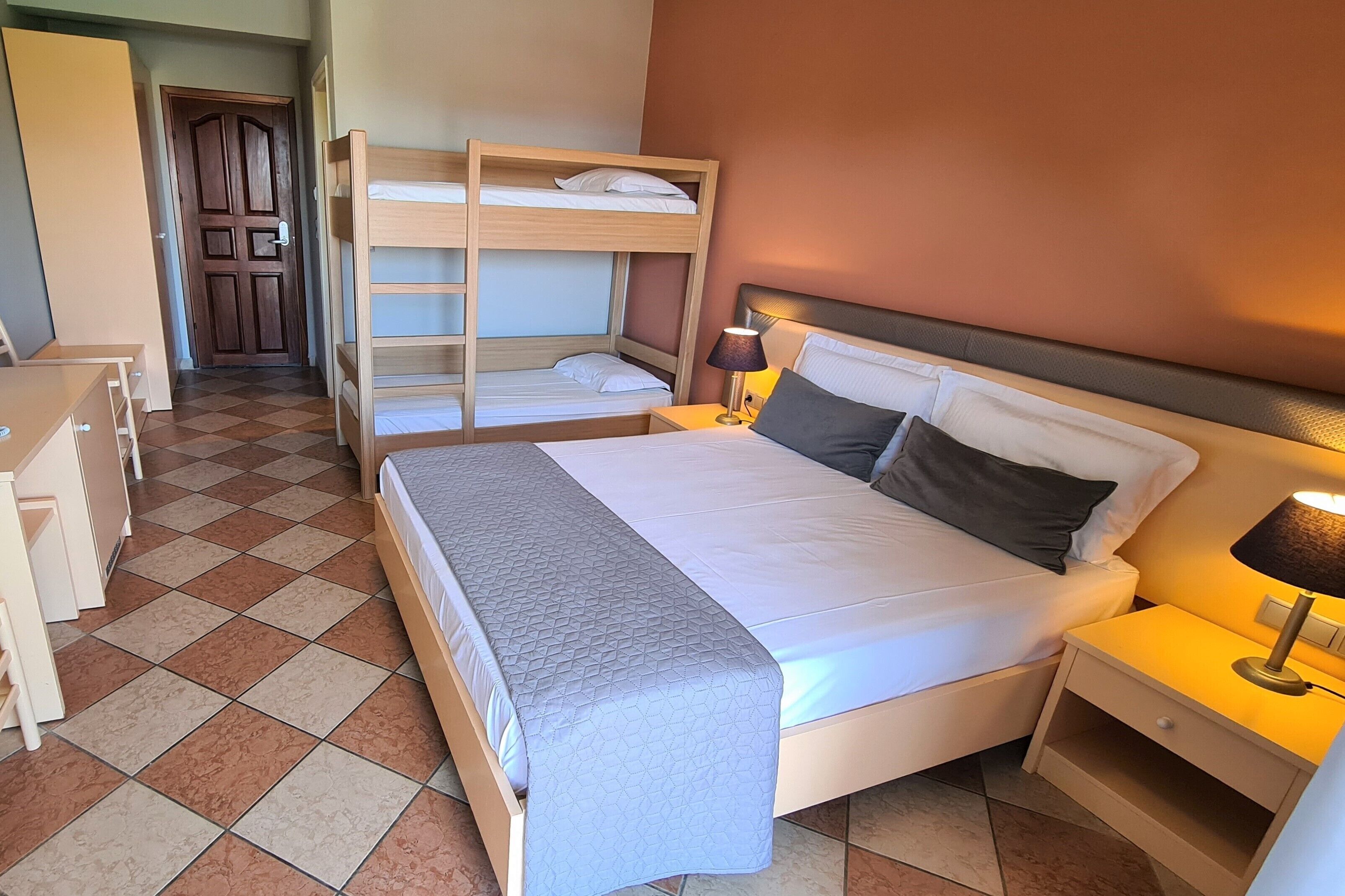 Ein Hotelzimmer mit einem Etagenbett, einem Einzelbett, einem Schreibtisch und einem Nachttisch mit Lampe.