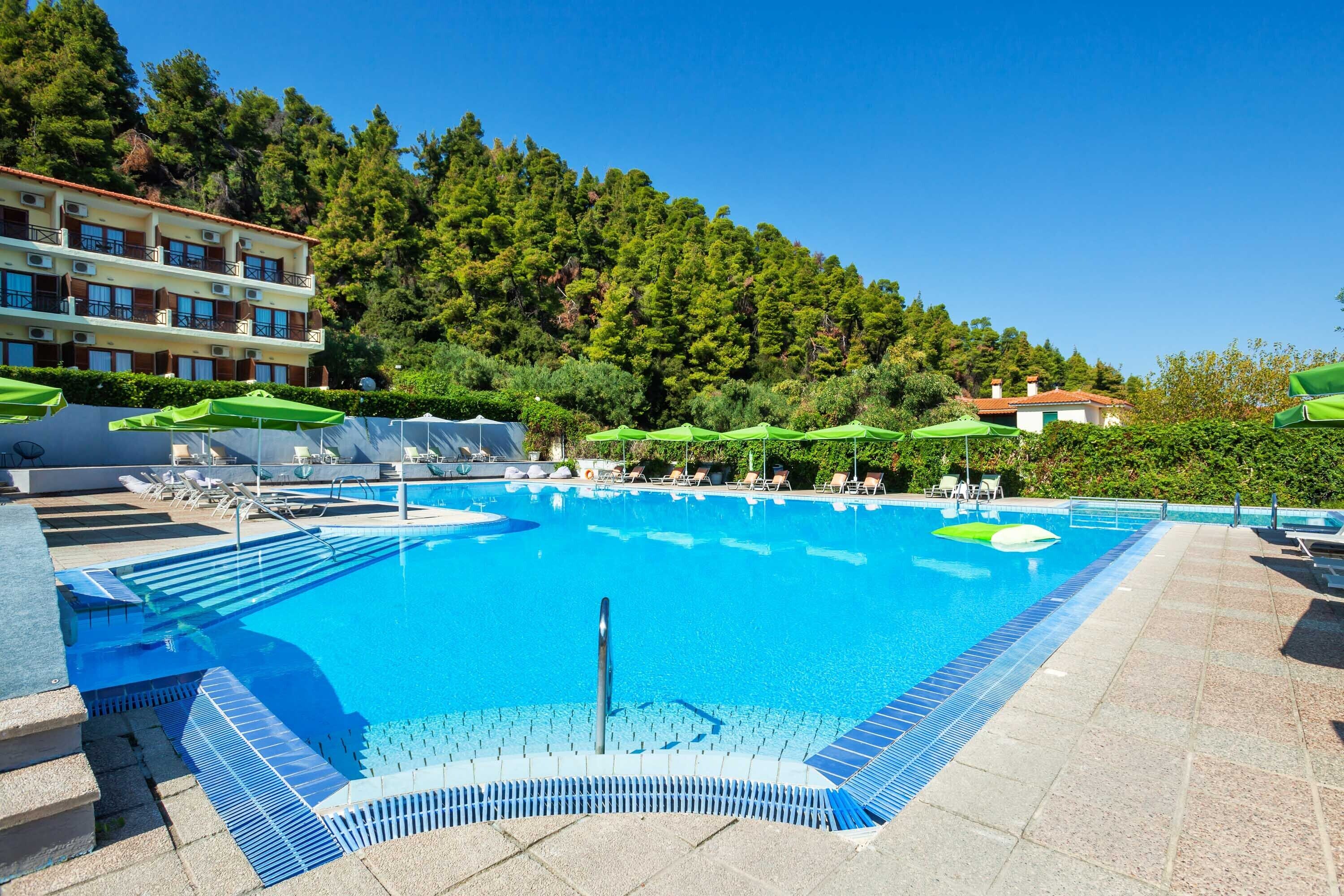 A swimming pool with lounge chairs and umbrellas, surrounded by a building and trees.