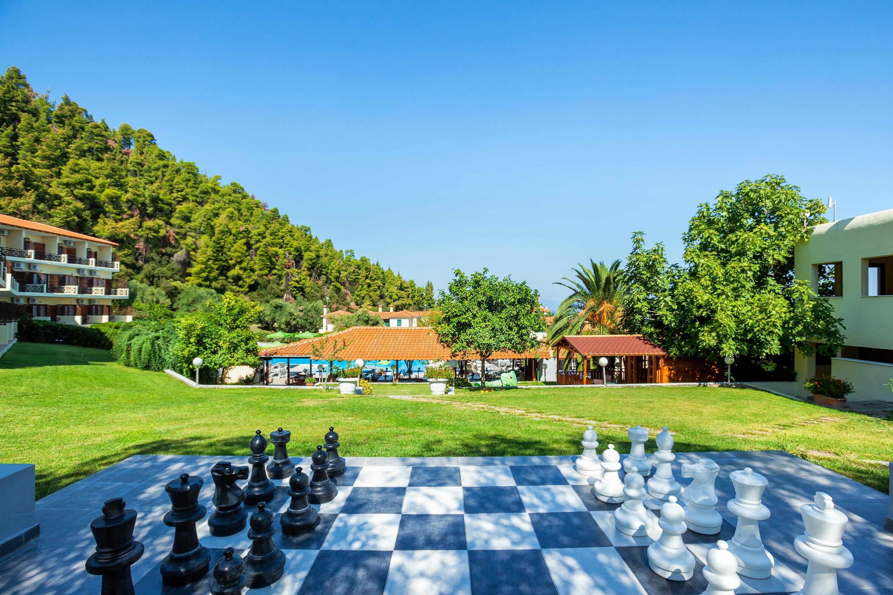 A large outdoor chessboard set up on a grassy area with buildings in the background.