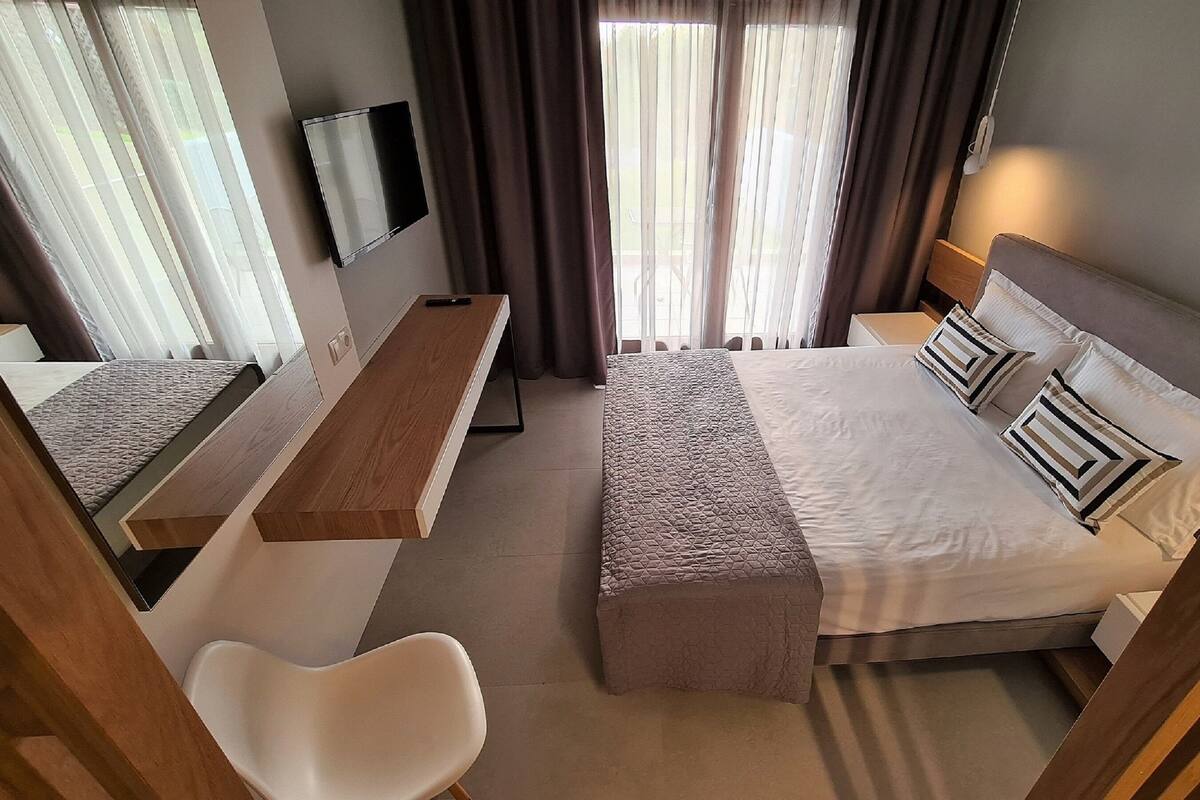 Ein Hotelzimmer mit Bett, Schreibtisch, Stuhl, Fernseher und einem Fenster mit Vorhängen.