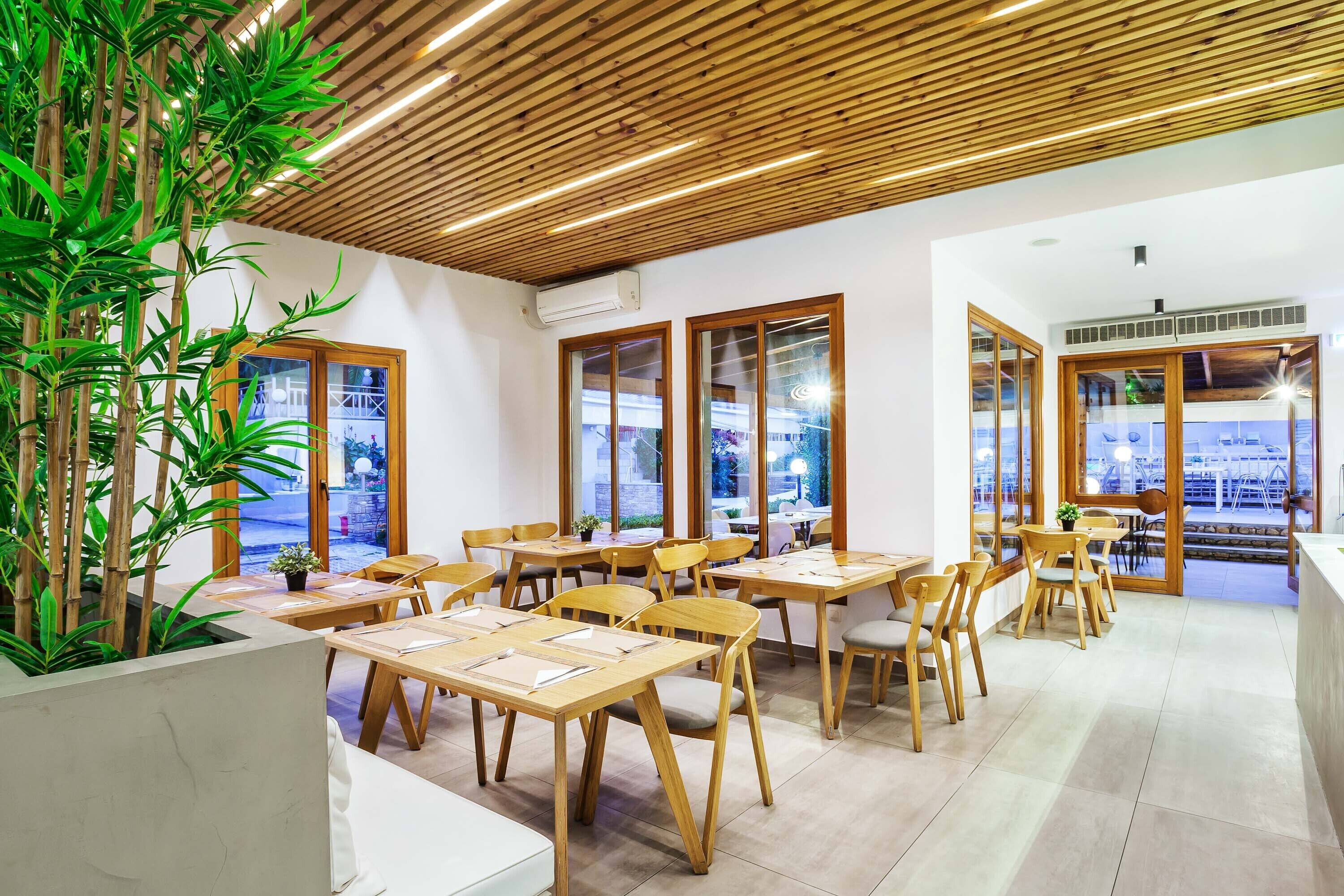 Un espace repas moderne avec des tables et des chaises en bois, de grandes fenêtres et des plantes d’intérieur.