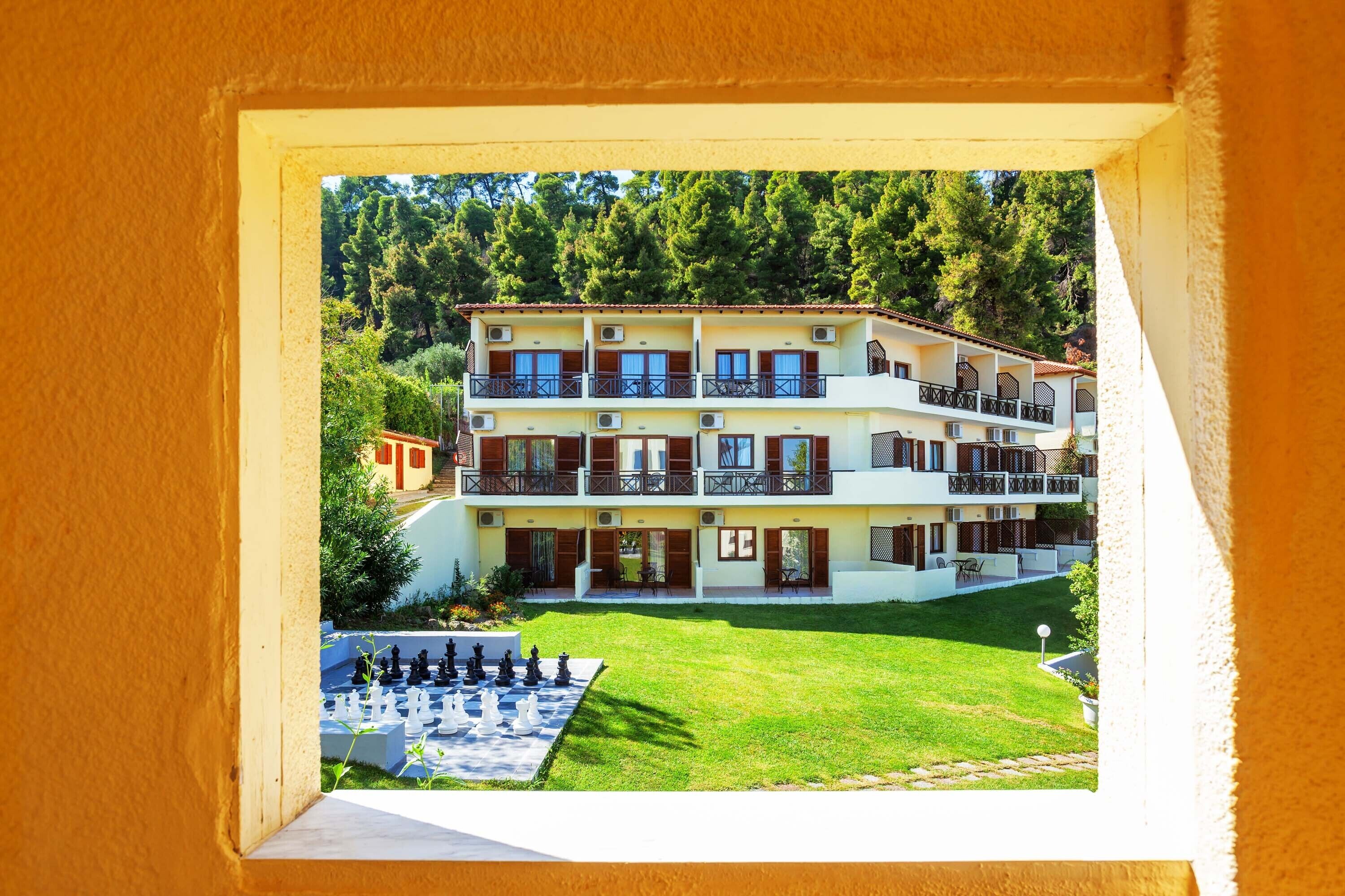 Un edificio de varios pisos con balcones, un tablero de ajedrez en primer plano y un paisaje verde exuberante al fondo.