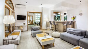 A modern living room with a sofa, ottoman, and coffee table, a dining area with chairs, and a kitchen with bar stools. - Palladium Hotel (Kassandra)