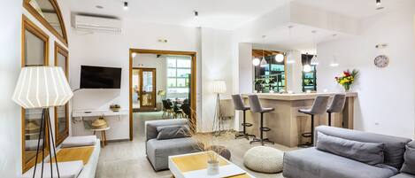 A modern living room with a sofa, ottoman, and coffee table, a dining area with chairs, and a kitchen with bar stools.