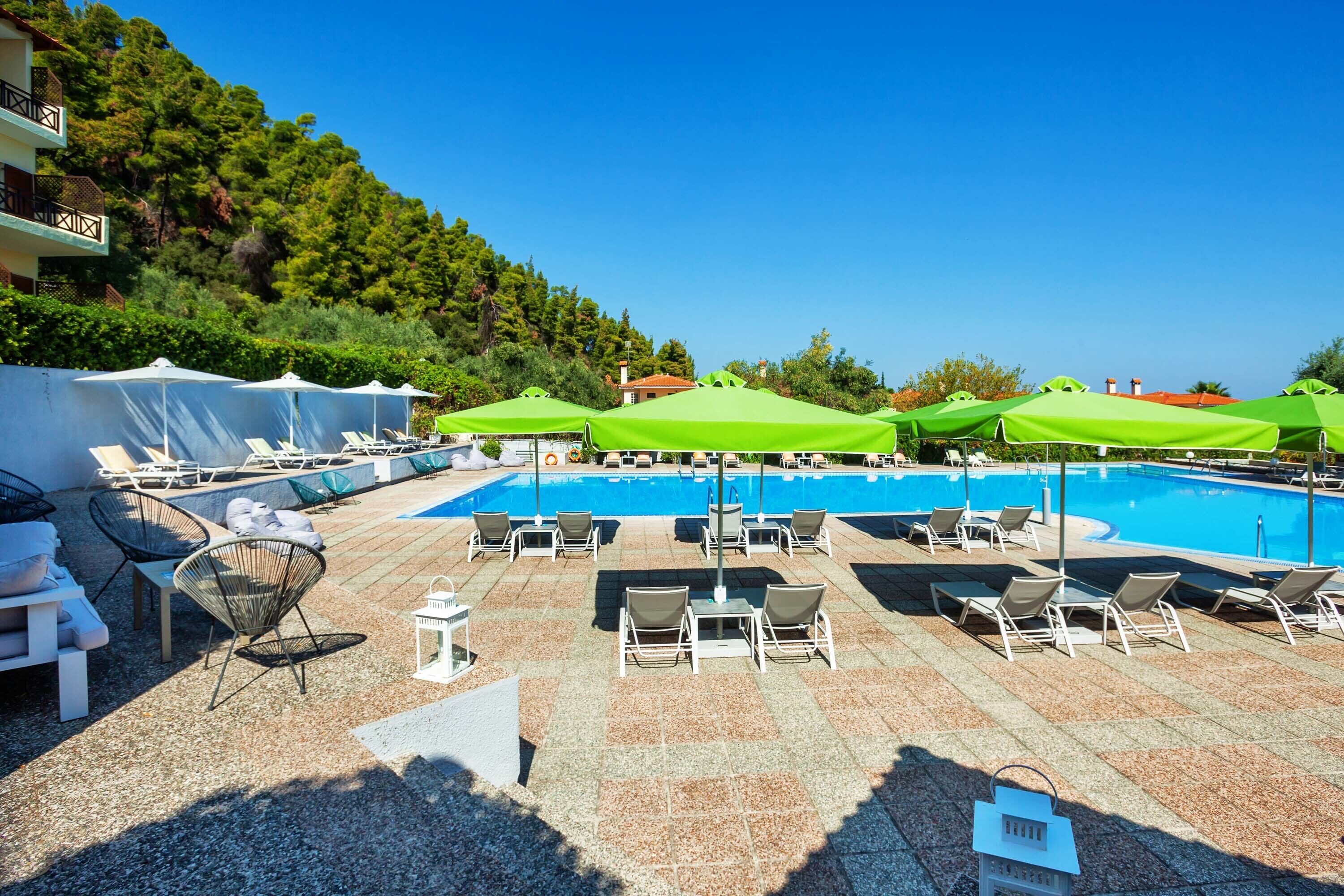 Zona de piscina con sillones de descanso y sombrillas, rodeada por un suelo de piedra y un fondo de árboles y edificios.