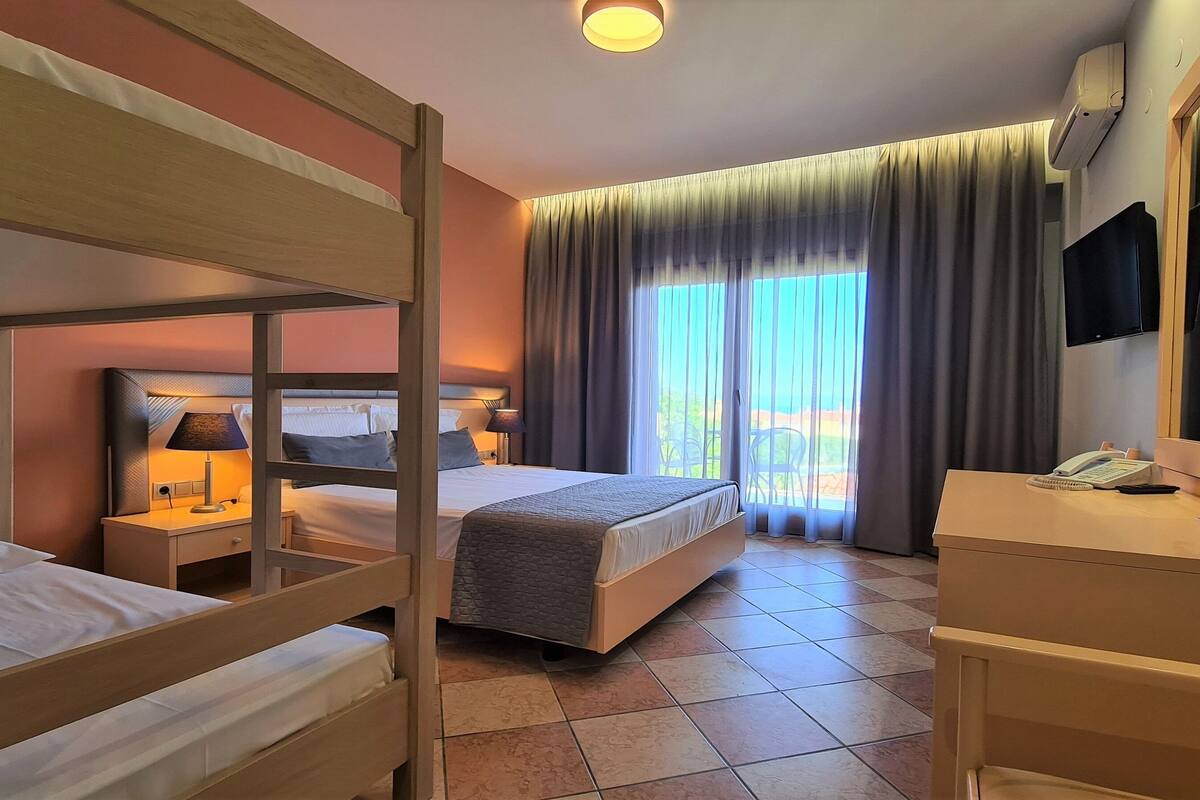 Ein Hotelzimmer mit Etagenbett, einem Einzelbett, einem Schreibtisch mit Telefon und einem großen Fenster mit Vorhängen.