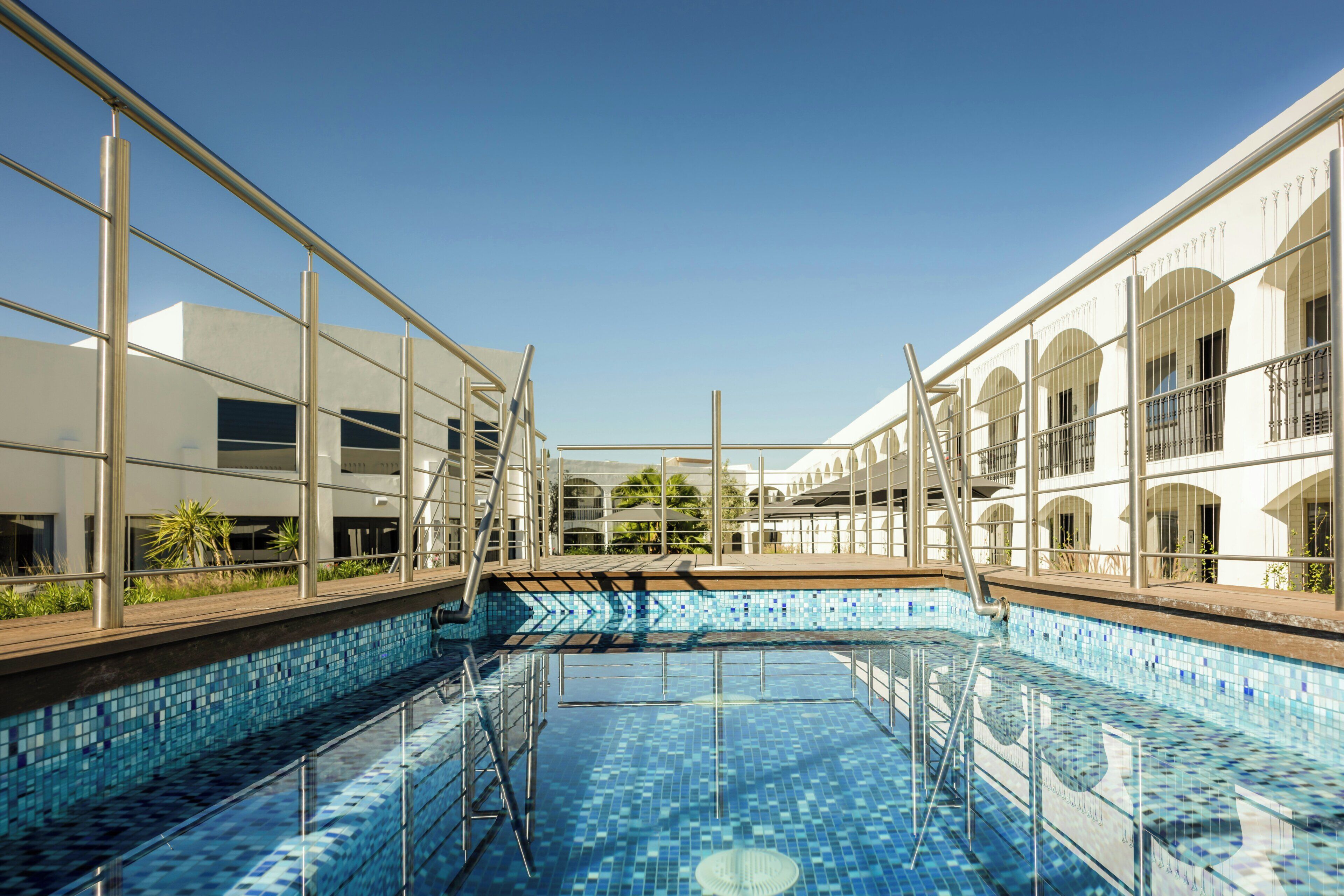 A swimming pool with clear blue tiles, surrounded by a metal railing, and adjacent to a building with large windows and balconies.