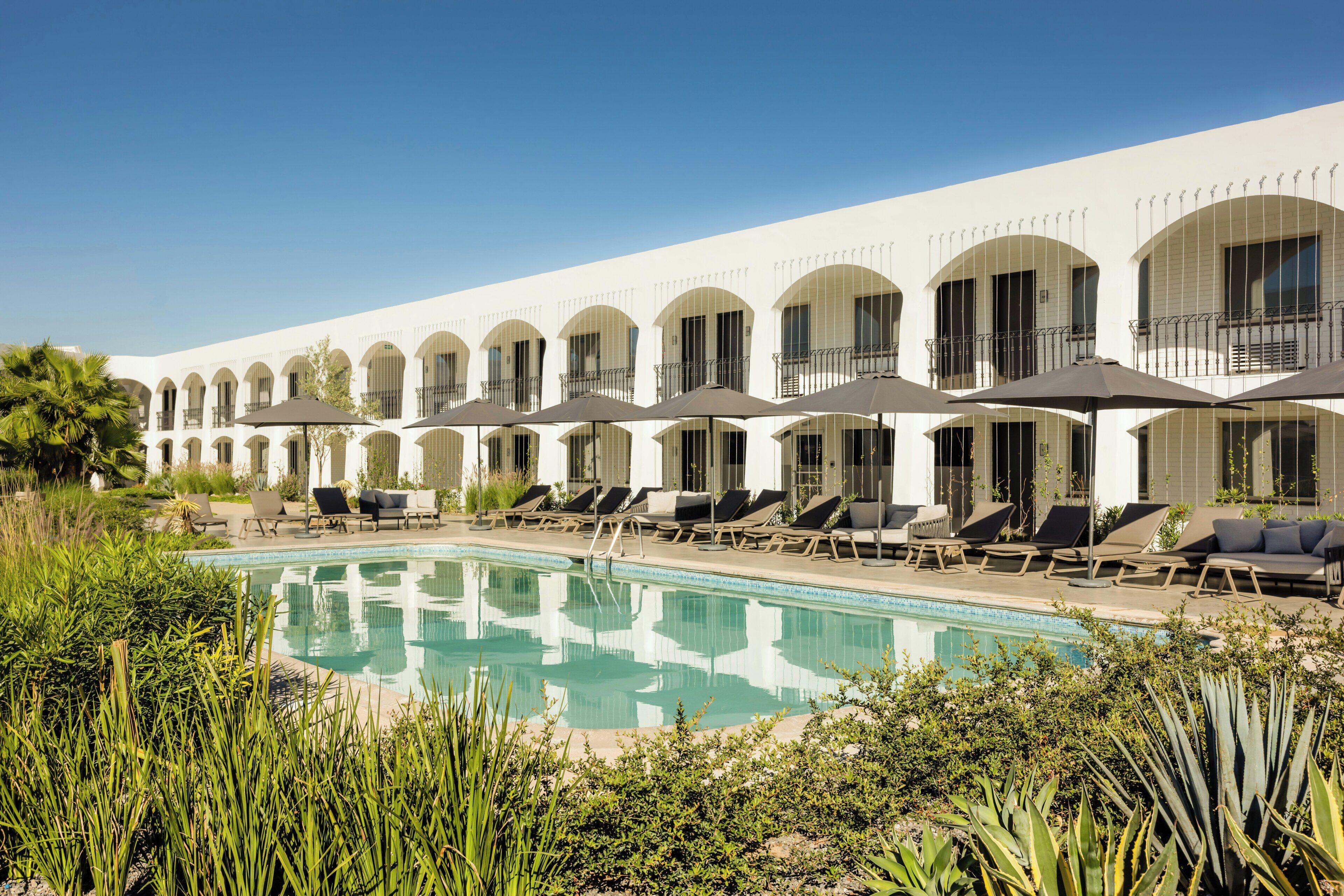 Un hôtel doté d’une piscine, de chaises longues et d’un bâtiment blanc aux fenêtres en arc de cercle.