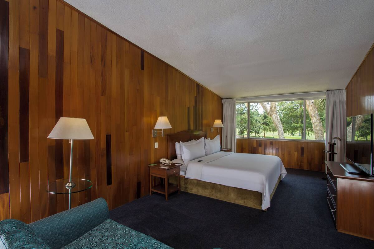 A hotel room with a bed, two bedside lamps, a television, a small table, and a sofa.