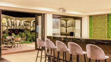 A modern bar area with a black countertop and four bar stools. There is a glass partition with a view of a courtyard with arches and greenery.