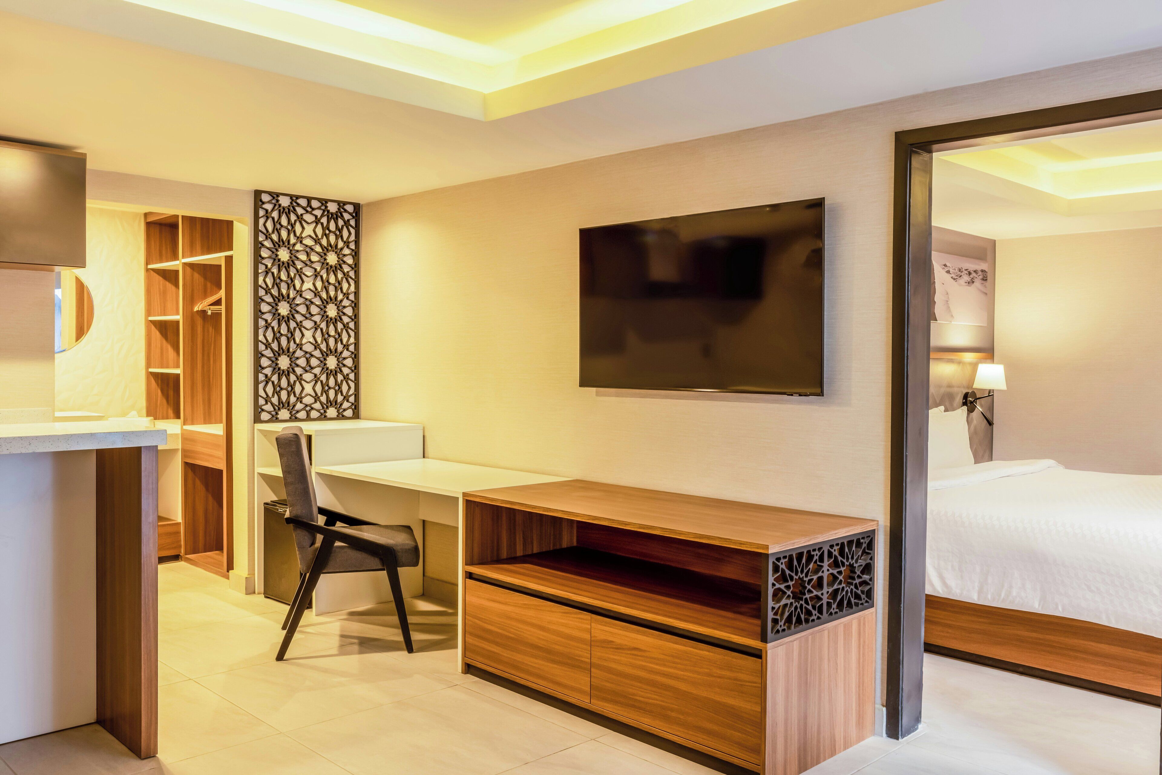 A modern hotel room with a wooden desk, a TV mounted on the wall, a bed with white linens, and a walk-in closet.