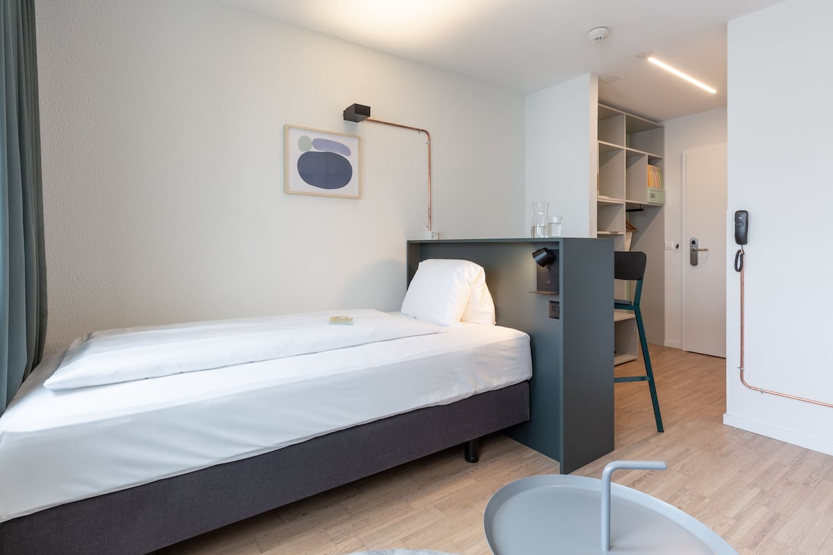 Ein ordentlich bezogenes Bett mit weißen Leinen, ein Schreibtisch mit Stuhl und ein Regal in einem modernen Zimmer mit hellem Holzboden.