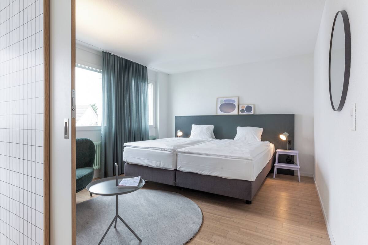 Ein modernes Hotelzimmer mit Doppelbett, Nachttischen, einem kleinen runden Tisch und Blick ins Freie.