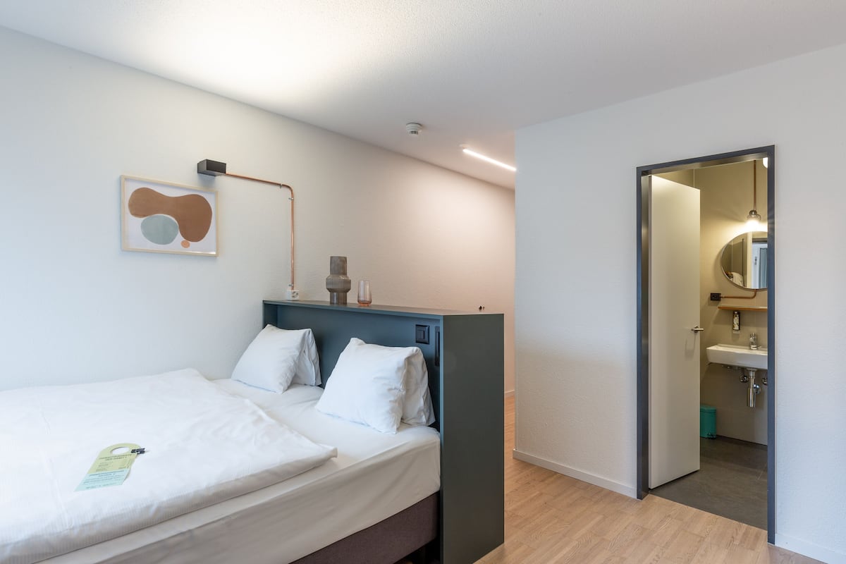 Ein Hotelzimmer mit einem Bett, einem Nachttisch, einem Spiegel und einem durch eine offene Tür einsehbaren Badezimmer.