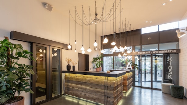 A modern reception area with a wooden reception desk, hanging pendant lights, and a glass door leading to a winter garden. - Alpenblick Bern (Bern)