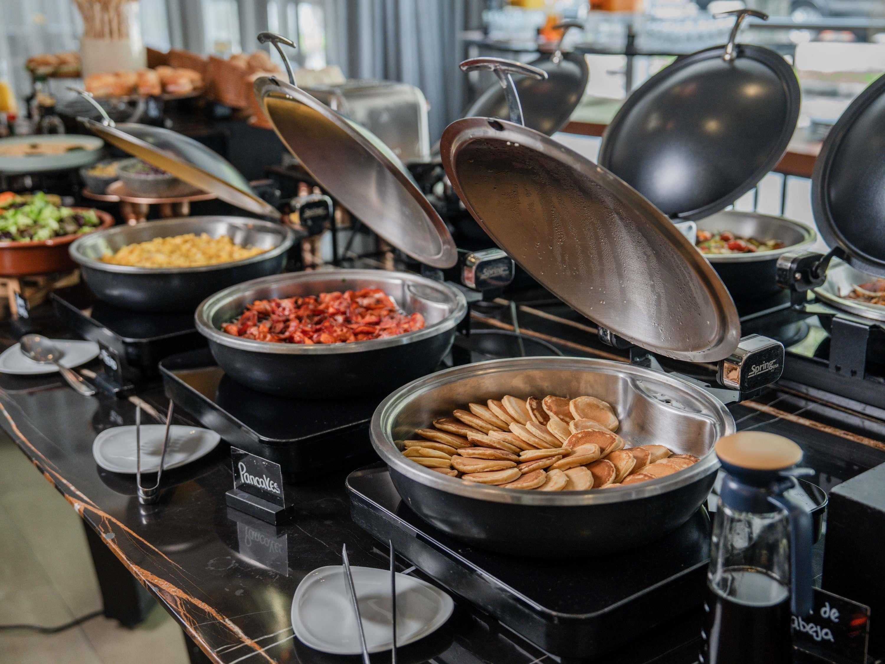 A buffet setup with various food items in bowls and pans, including pancakes and a dish that appears to be a type of casserole or salad.