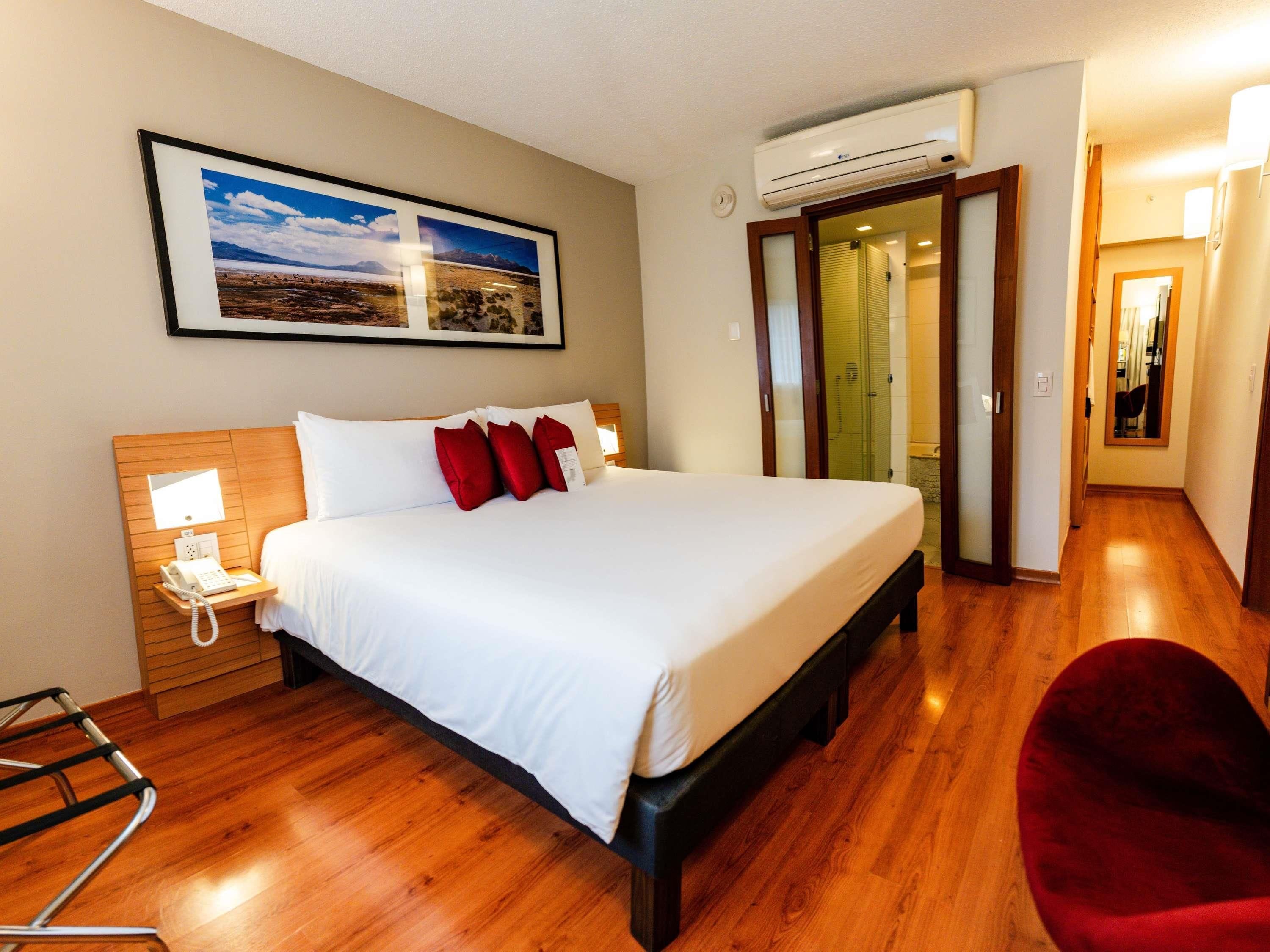 A hotel room with a bed, a bedside phone, a red chair, and a framed picture on the wall.