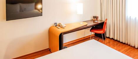 A hotel room with a bed, a desk with a telephone, a chair, a TV mounted on the wall, and a window with curtains.