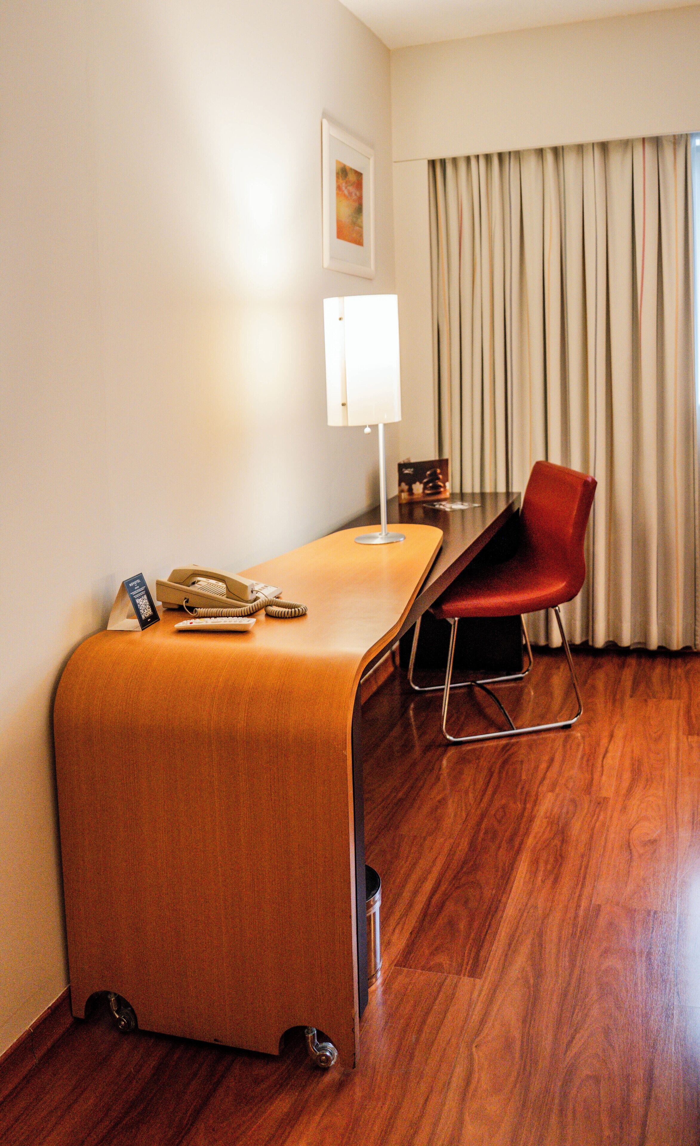 A hotel room with a wooden desk, a red chair, a lamp, and a telephone.