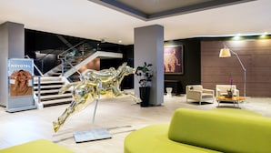 A modern hotel lobby with a central staircase, a Novotel sign, a metallic horse sculpture, and green seating areas.