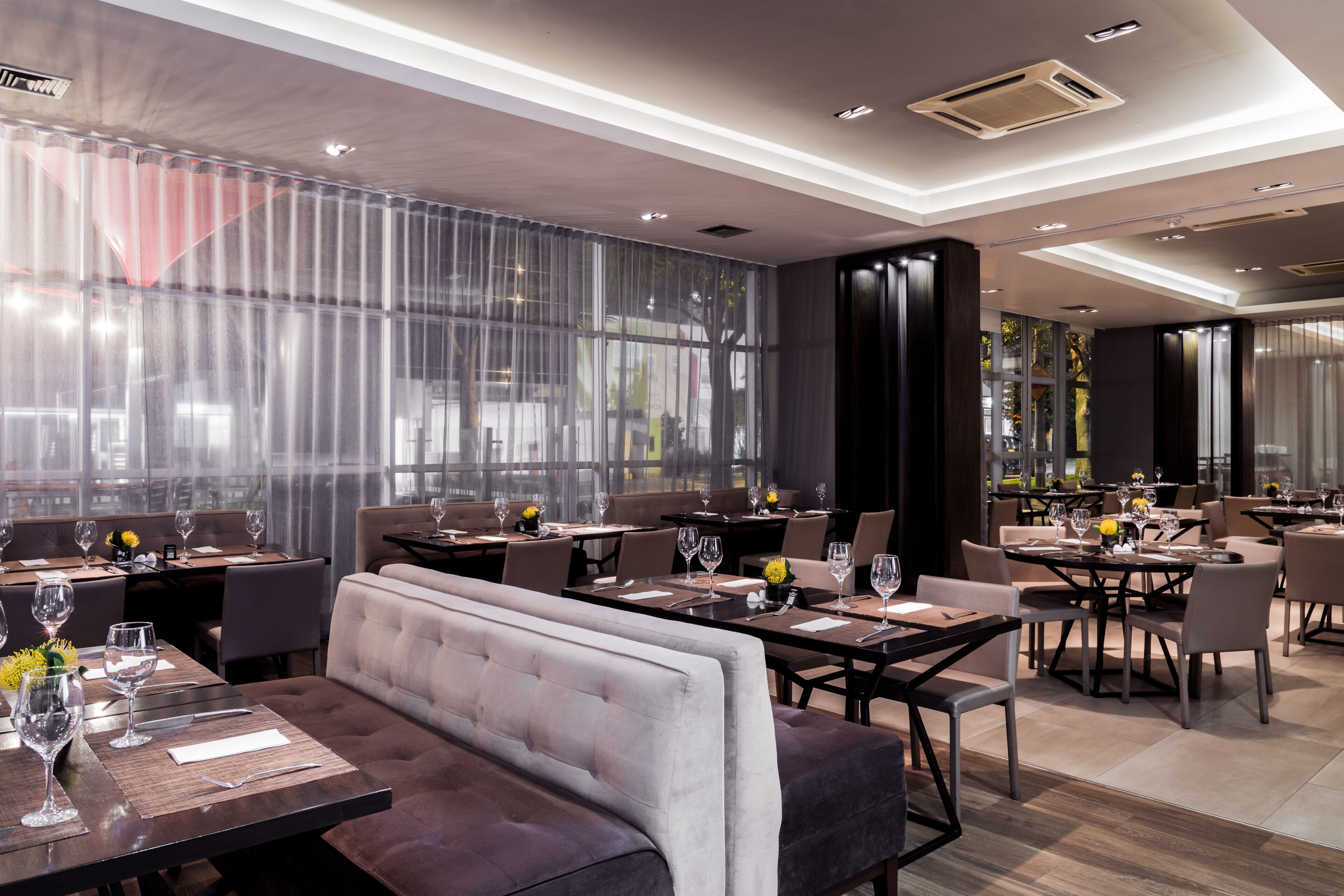Un restaurant moderne doté de hauts plafonds, de grandes fenêtres et d’un agencement varié des espaces, comprenant des tables et des banquettes.
