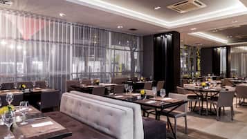 Un restaurant moderne doté de hauts plafonds, de grandes fenêtres et d’un agencement varié des espaces, comprenant des tables et des banquettes.