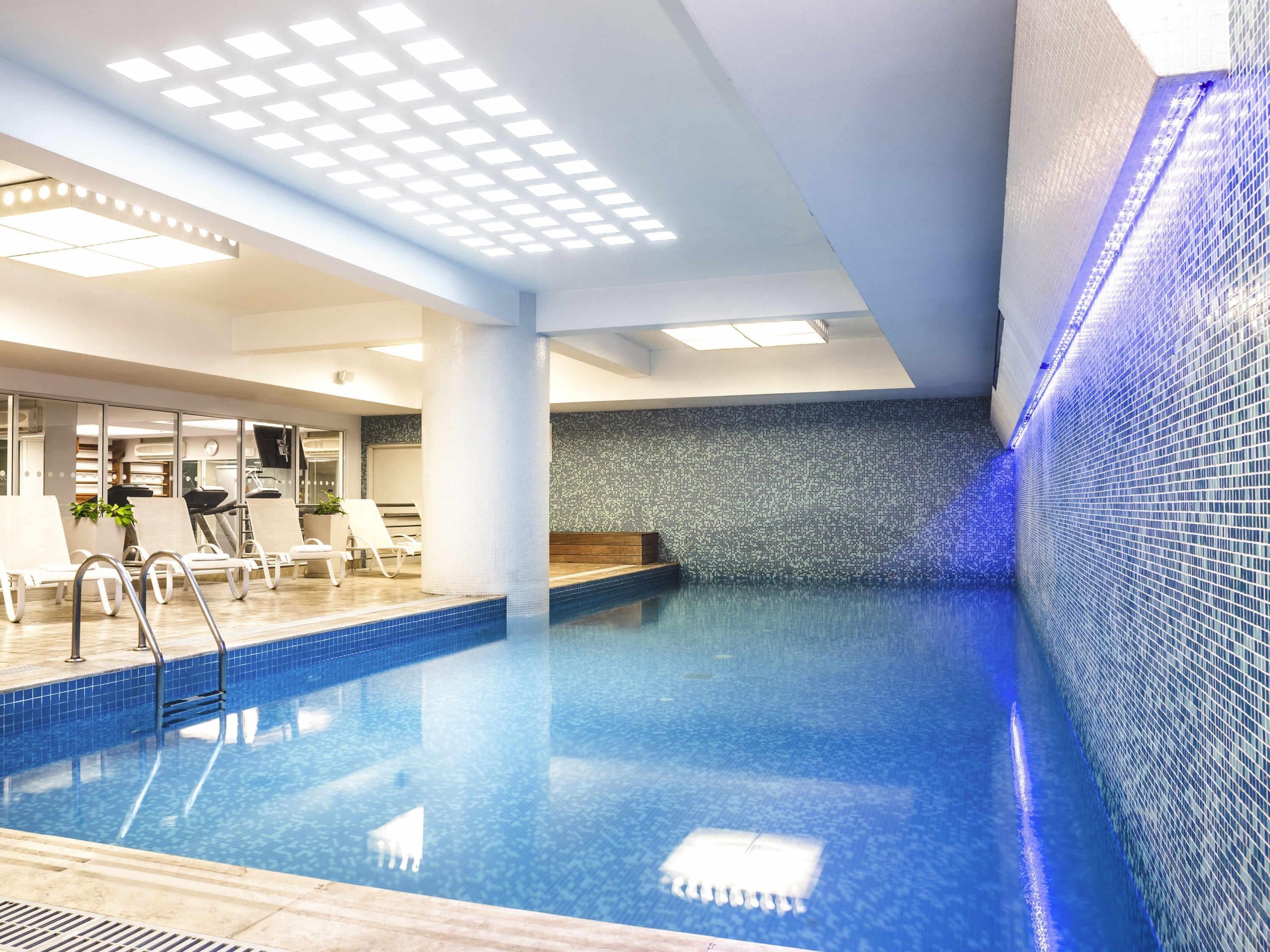 Una piscina interior con pared de azulejos, techo con iluminación empotrada y una zona de estar con sillas y una planta.
