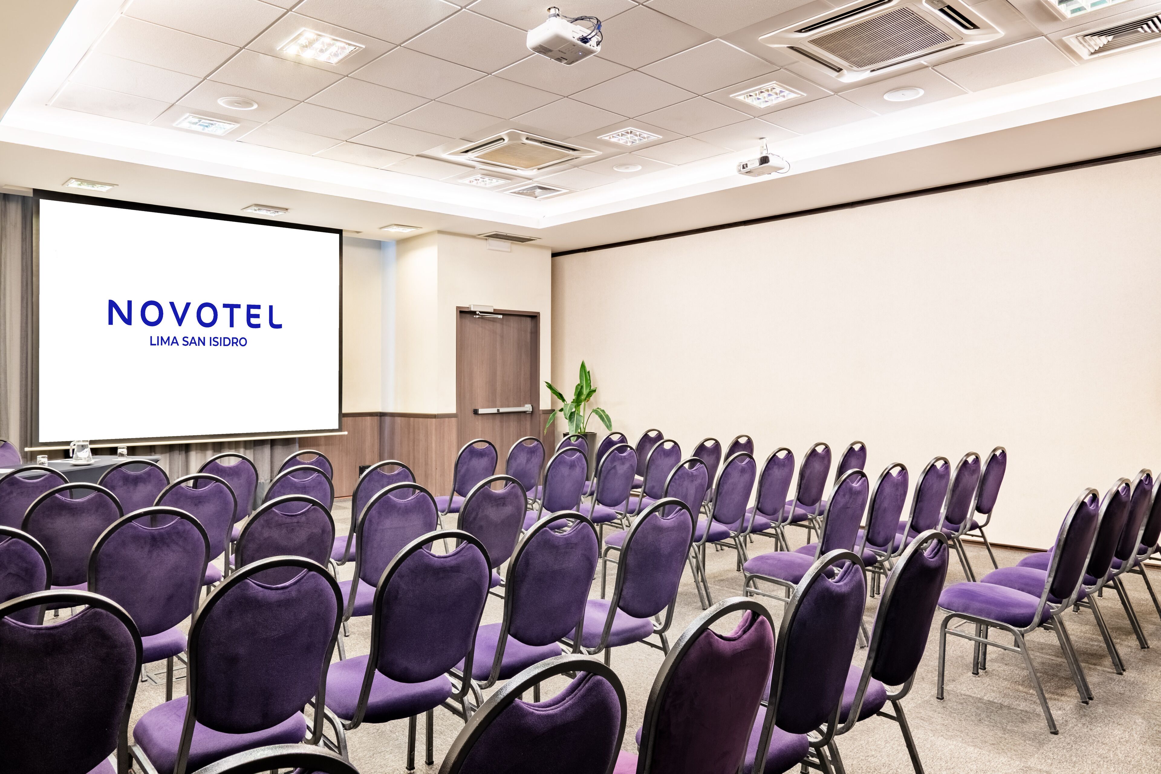 Konferenssisali, jossa on riveissä violetteja tuoleja, projektorikangas, jossa näkyy teksti ”NOVOTEL LIMA SAN ISIDRO”, ja taustalla ovi.