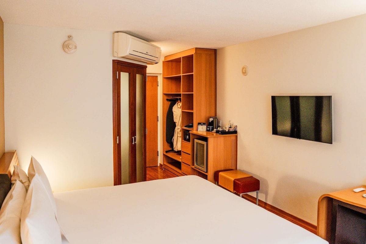 A hotel room with a bed, a TV, a minibar, a wardrobe, and an air conditioning unit.
