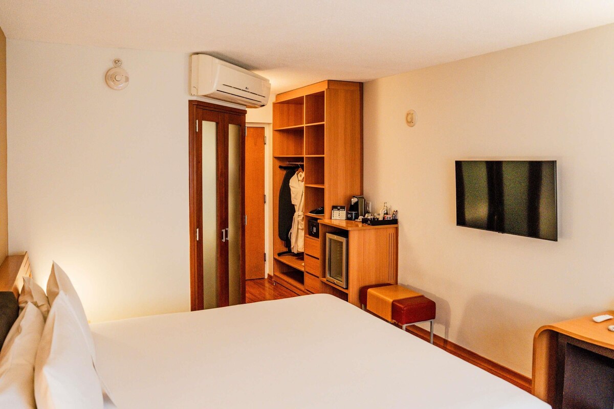 A hotel room with a bed, a TV, a minibar, a wardrobe, and an air conditioning unit.
