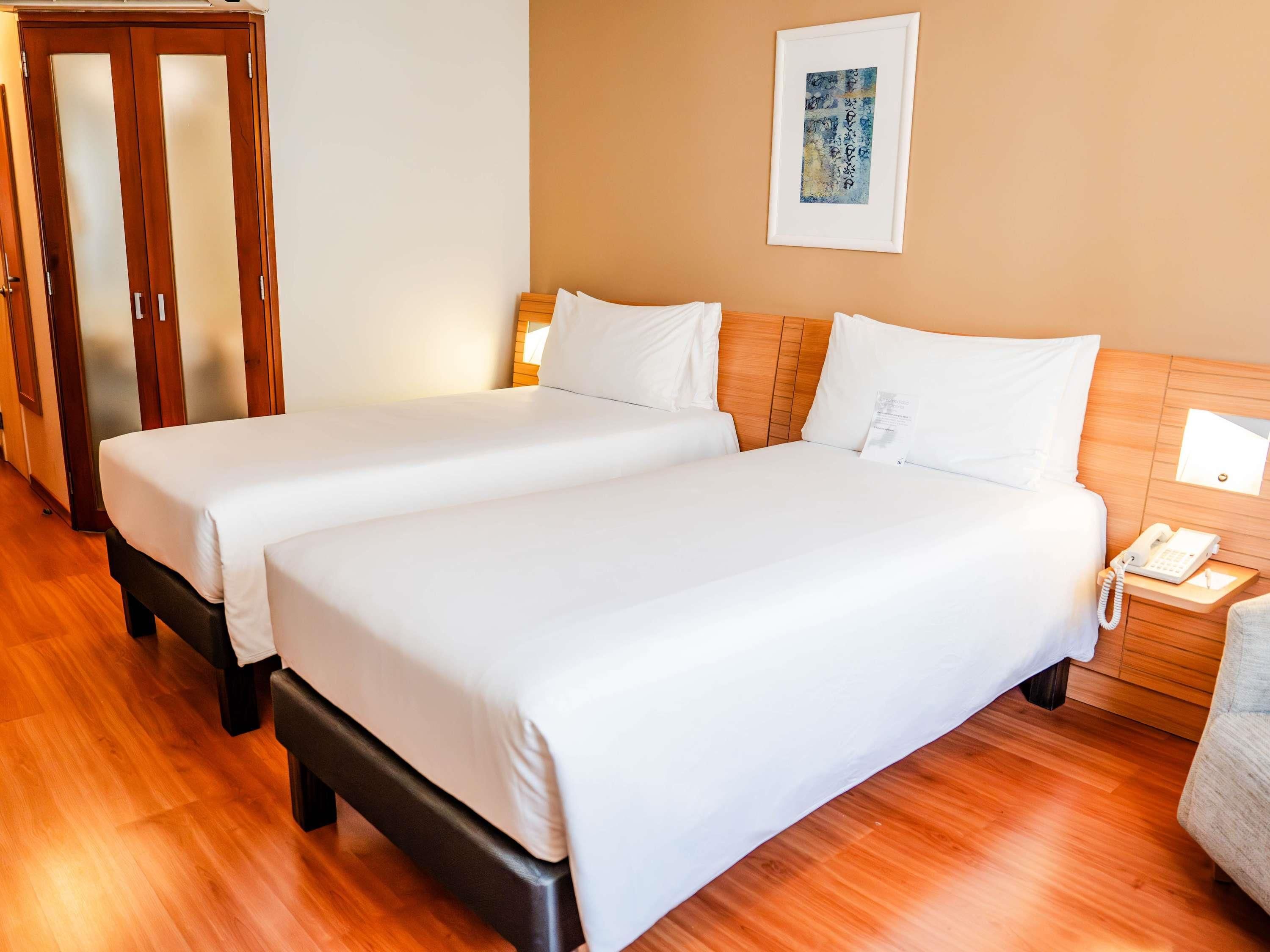 A hotel room with two beds, wooden flooring, a wooden headboard, a bedside table with a phone, and a framed artwork on the wall.