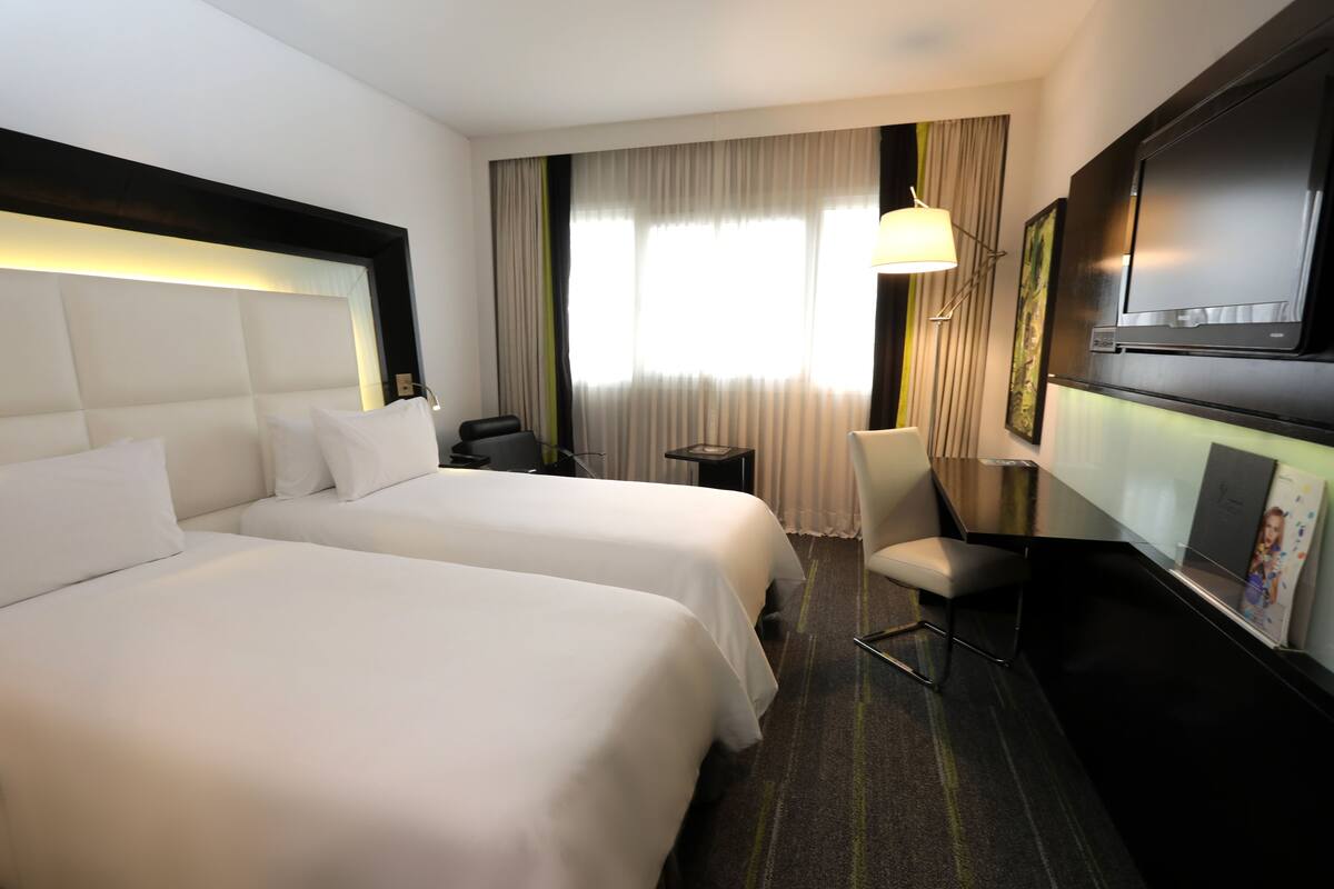 A hotel room with two beds, a desk, a chair, a TV, and a window with curtains.